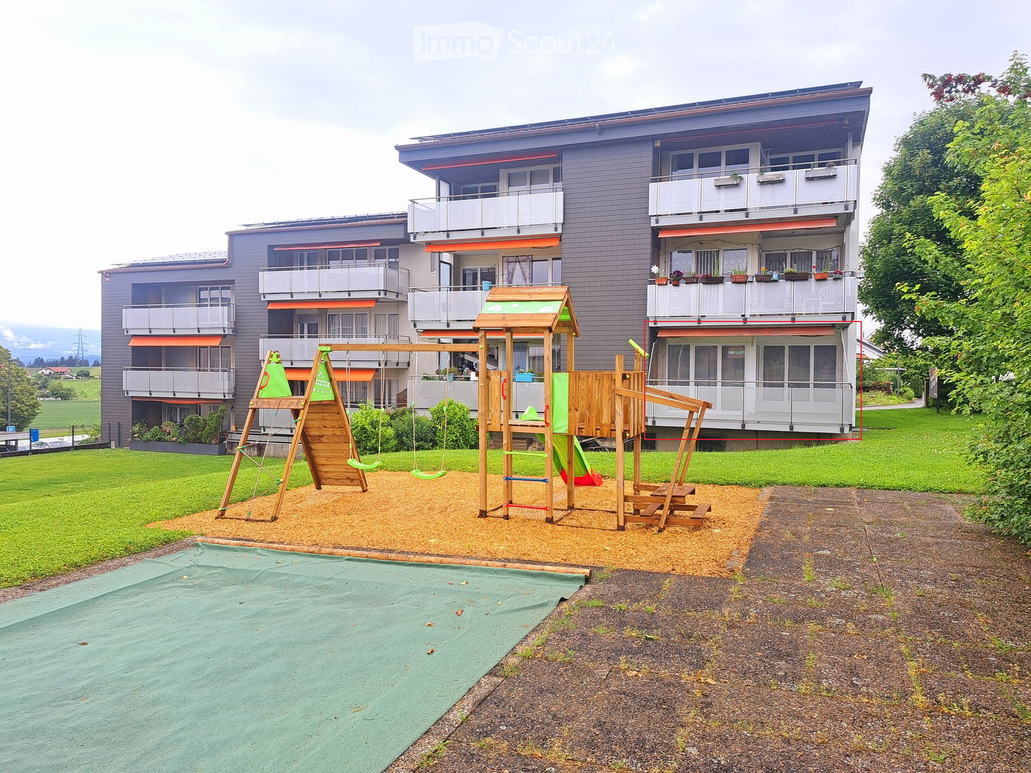 A multi-story residential building with balconies and a playground area featuring swings, a slide, and climbing structures.