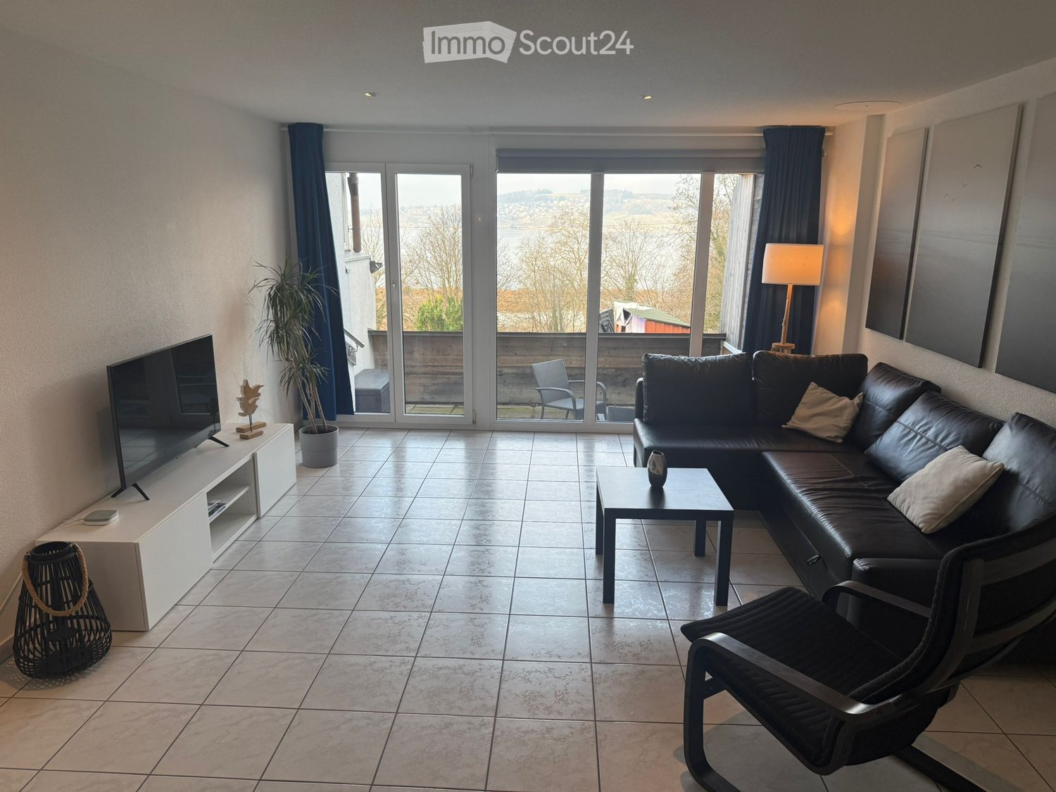 Modern living room with large windows offering a scenic view, featuring a black leather sofa, coffee table, armchair, TV, floor lamp, and decorative plant.