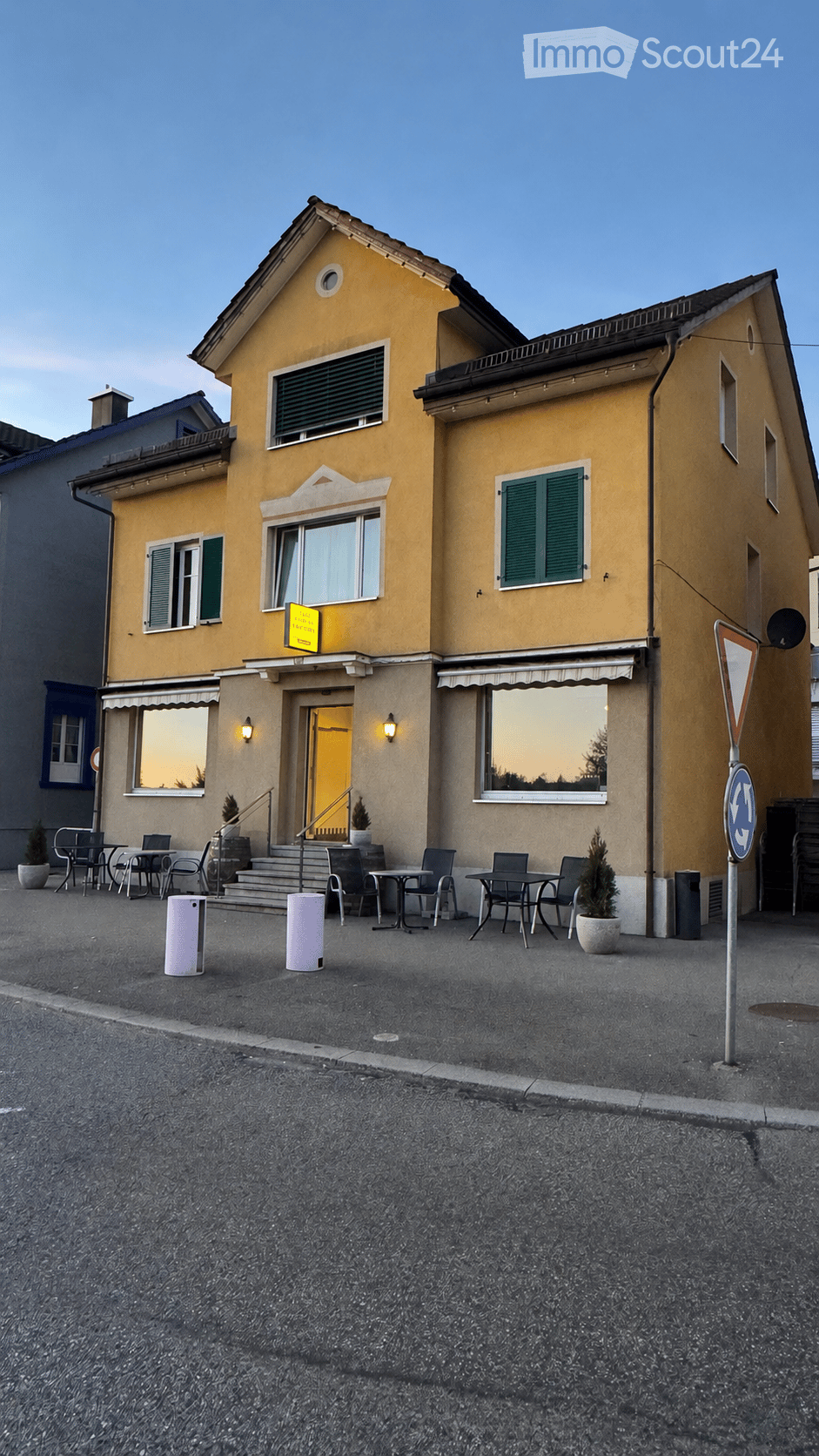 Two-story yellow building with green shutters, outdoor seating area with tables and chairs, small potted plants, and a signboard.