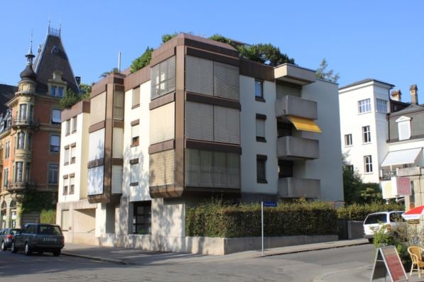 Modern multi-story apartment building with balconies and large windows, surrounded by other residential structures and parked cars.