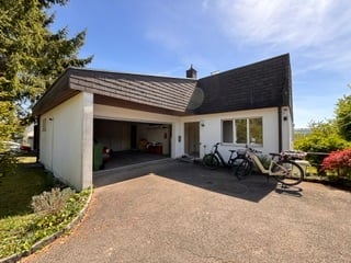 Modern single-story house with white exterior, dark grey roof, large garage door, and driveway with bicycles parked.