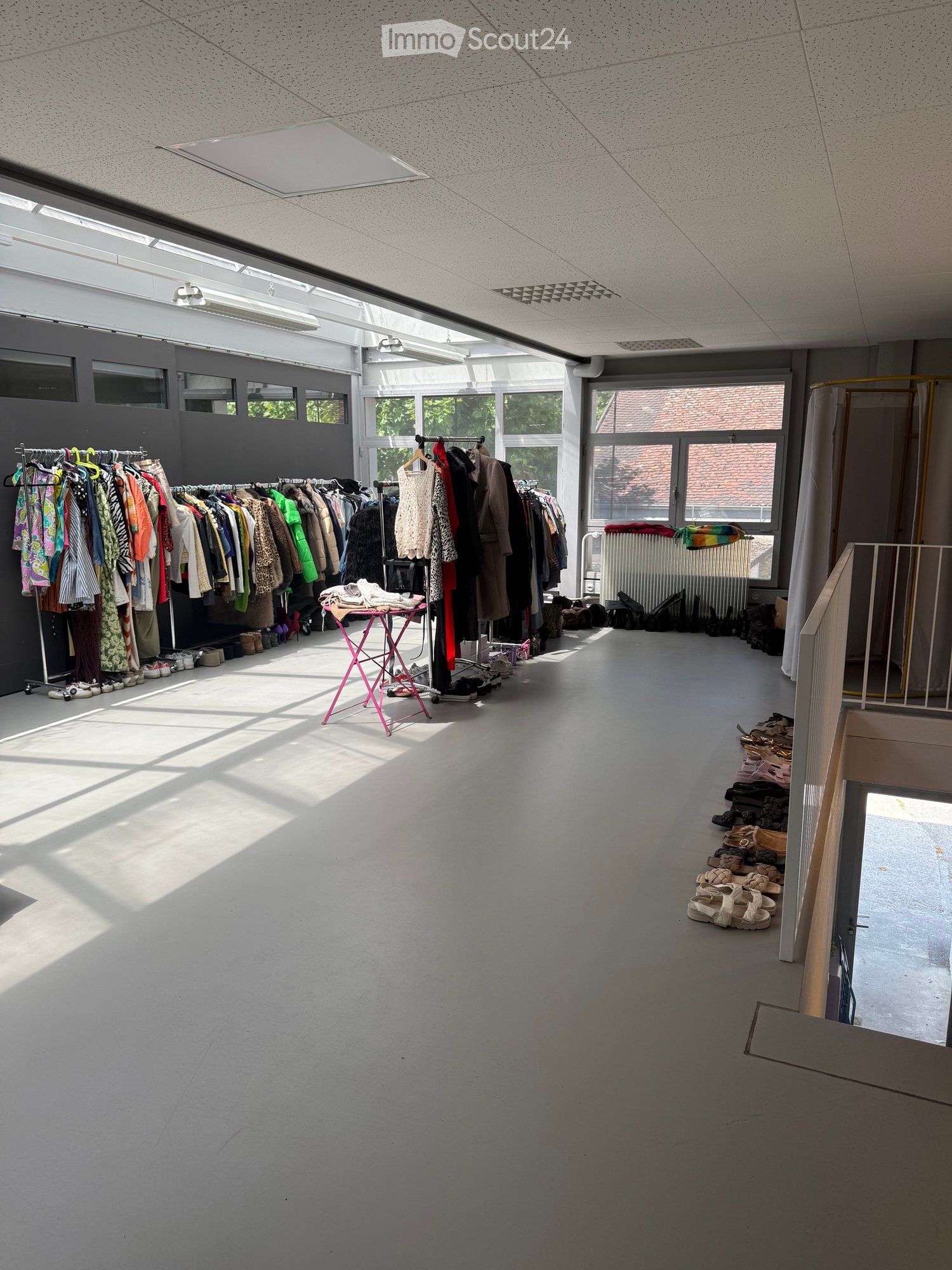 Bright, open room with large windows, clothing racks filled with garments, a small pink table, and shoes neatly arranged on the floor.