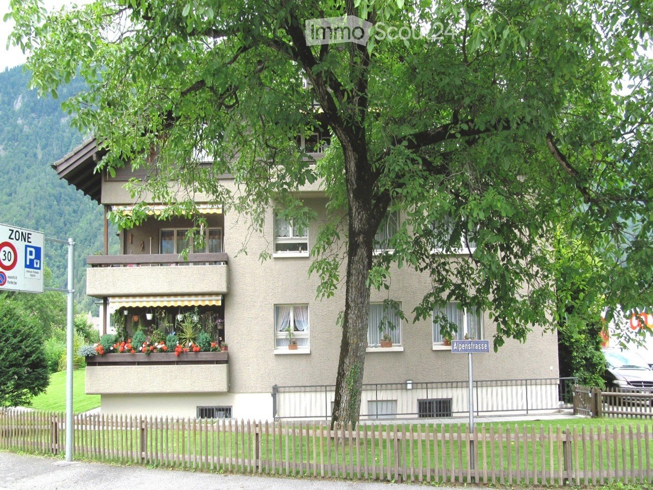 A two-story residential building with balconies, surrounded by greenery and a wooden fence.