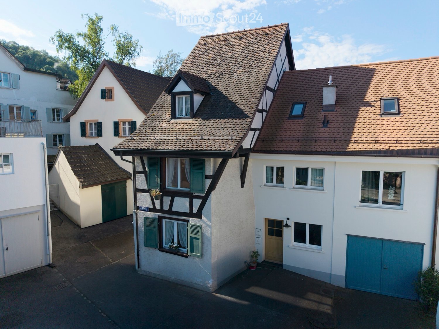 A cluster of traditional European houses with pitched roofs, white facades, and green shutters, featuring a central house with a prominent gabled roof and dormer windows.