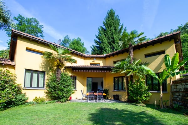 Two-story yellow house with a tiled roof, surrounded by lush greenery including palm trees and other vegetation. The house has a covered patio or balcony area, and a garage or parking space is visible.