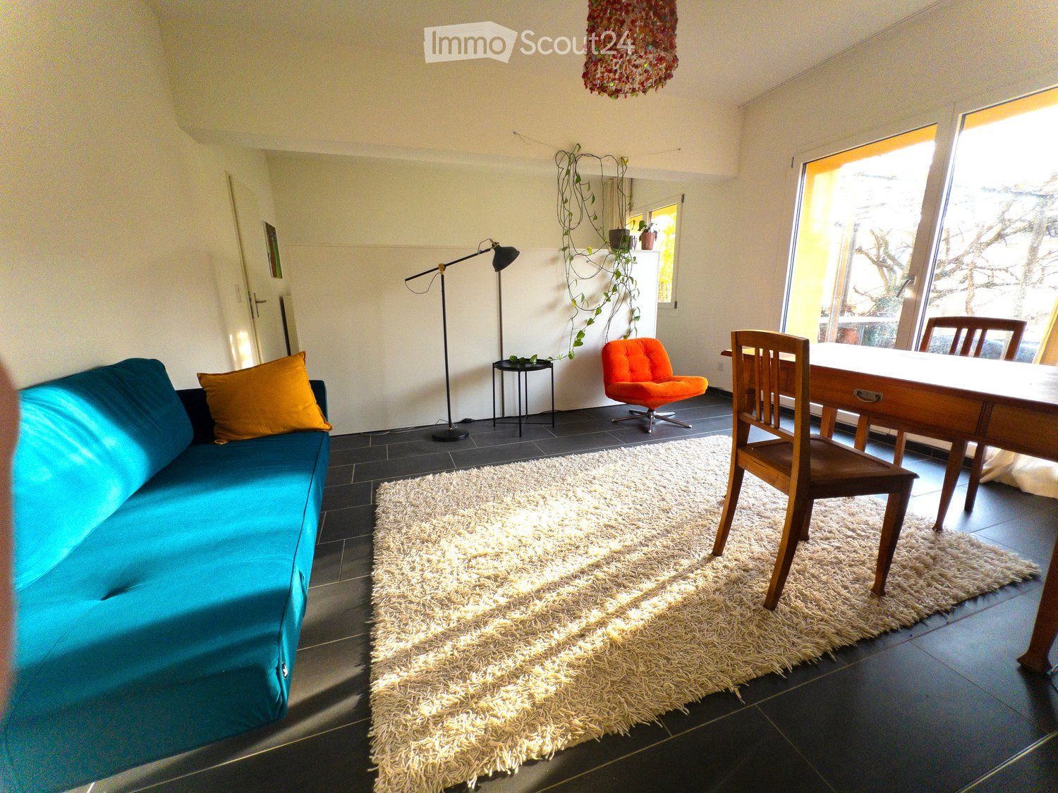 Bright living room with a blue sofa, orange accent chair, wooden dining table, and a fluffy white rug on a dark tiled floor.