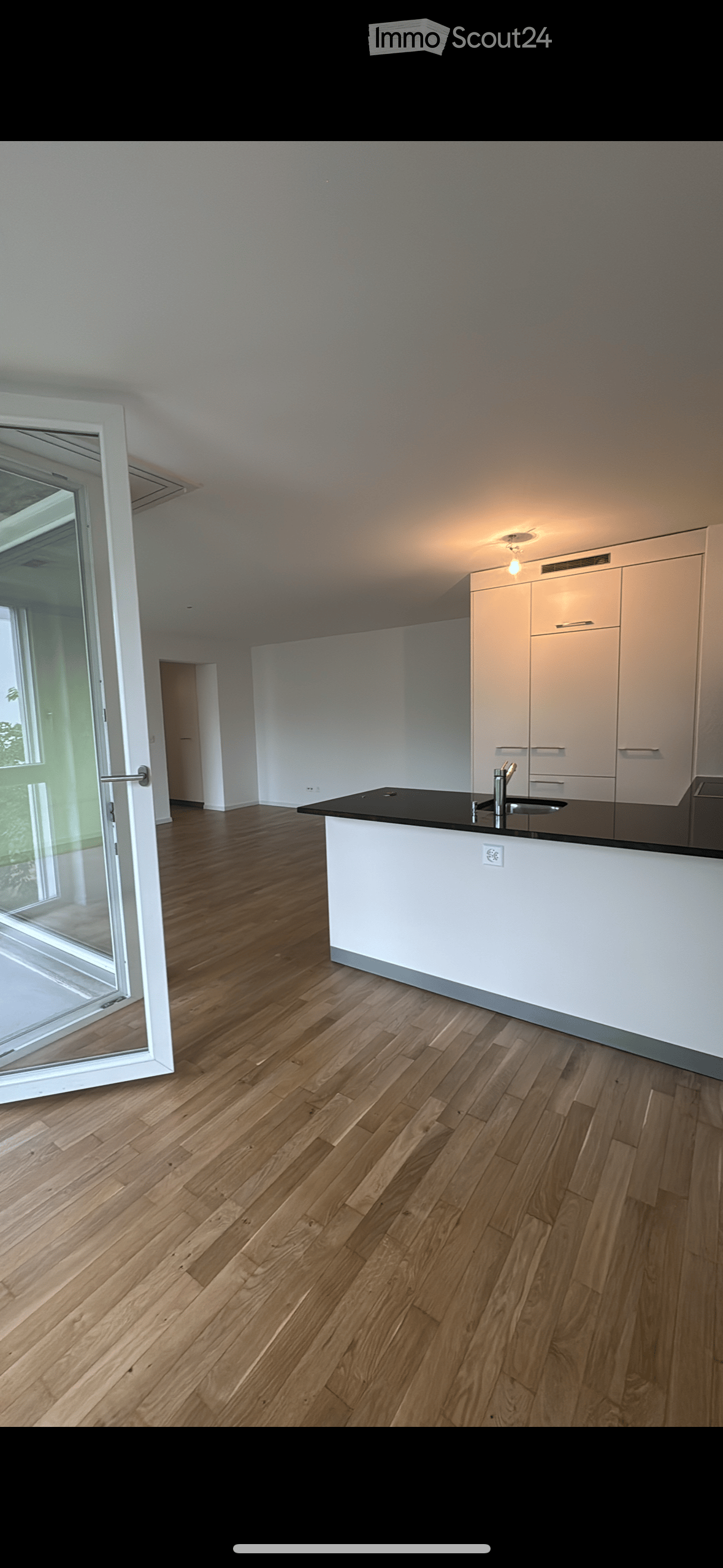 Modern, unfurnished kitchen with light wooden flooring, white cabinetry, and a black countertop. The space features large windows and a sliding glass door leading to an outdoor area.