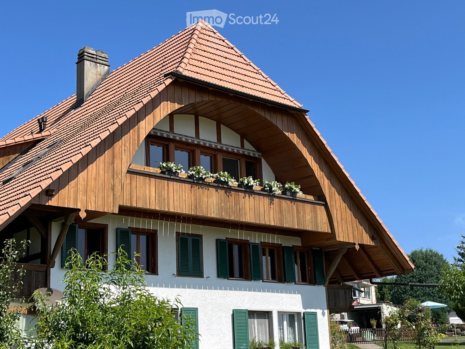 Two-story house with a wooden balcony, red-tiled roof, and green shutters. The balcony features potted plants, and the house is surrounded by greenery.