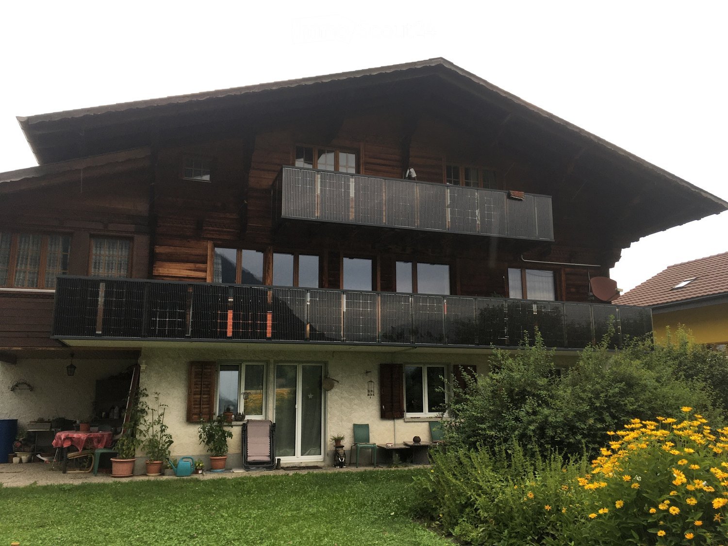 Two-story wooden house with large windows and balconies, surrounded by a well-maintained garden with potted plants and outdoor furniture.