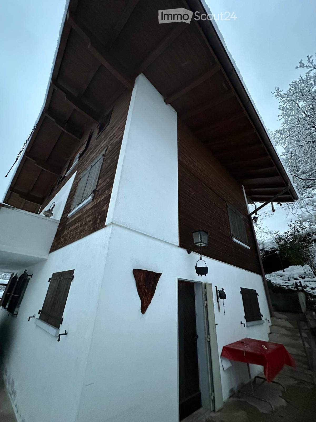Two-story house with white walls, wooden trim, and a slanted roof. Features include wooden shutters, a small porch with a table covered in a red cloth, and a hanging lantern.