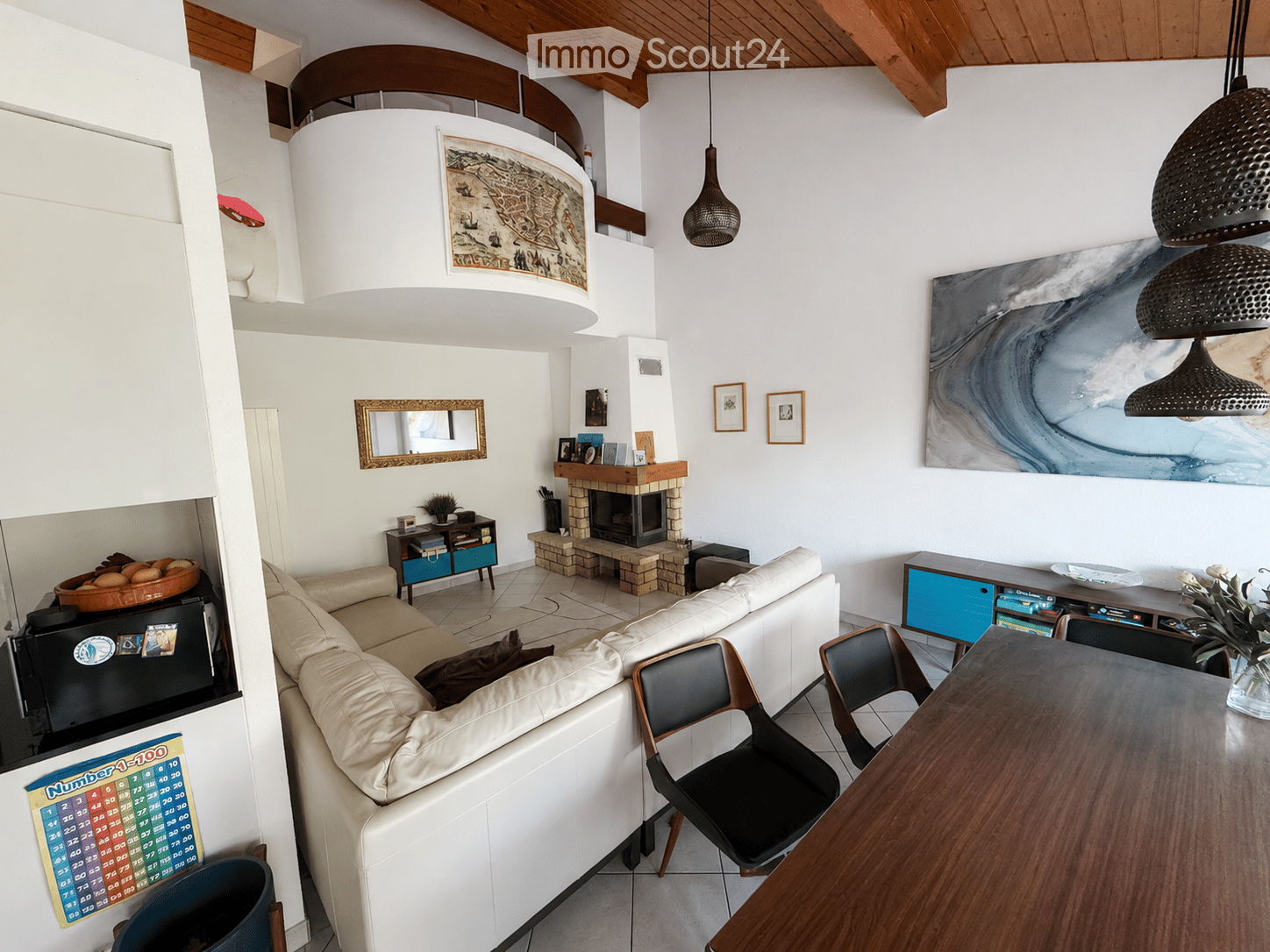 Modern living room with white walls, exposed wooden beams, a fireplace, a large sofa, a dining table with chairs, and decorative artwork.