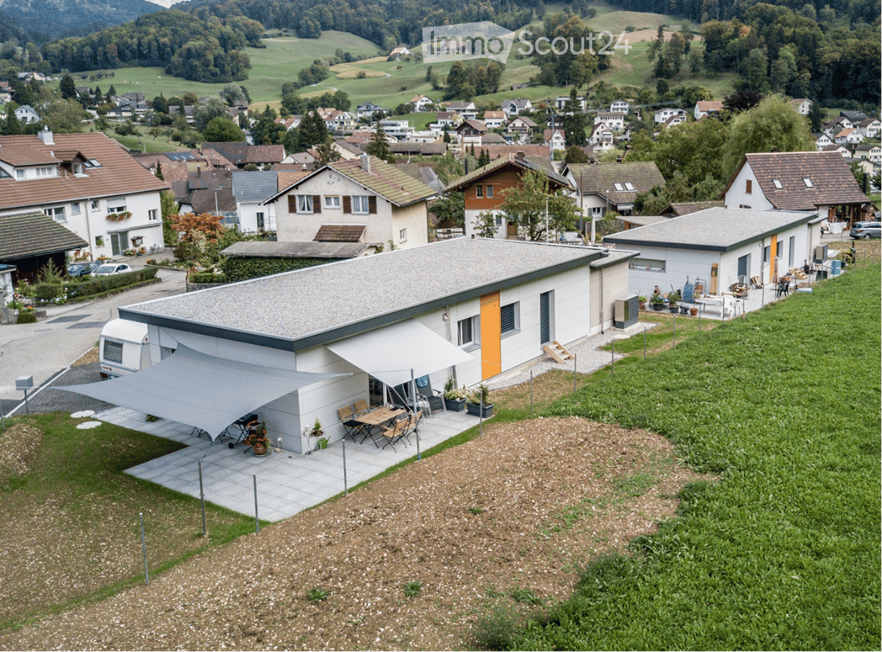 Modern single-story house with a grey roof and white walls, featuring a covered patio area with outdoor furniture and a scenic view of a village and green hills in the background.