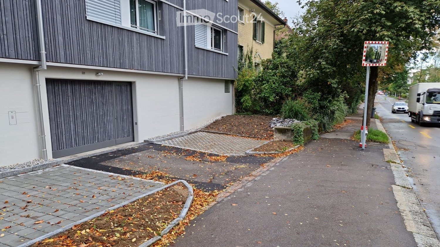 Two-story building with a garage, located on a paved driveway with autumn leaves scattered around. The building has a modern facade with grey siding and white trim around the windows.