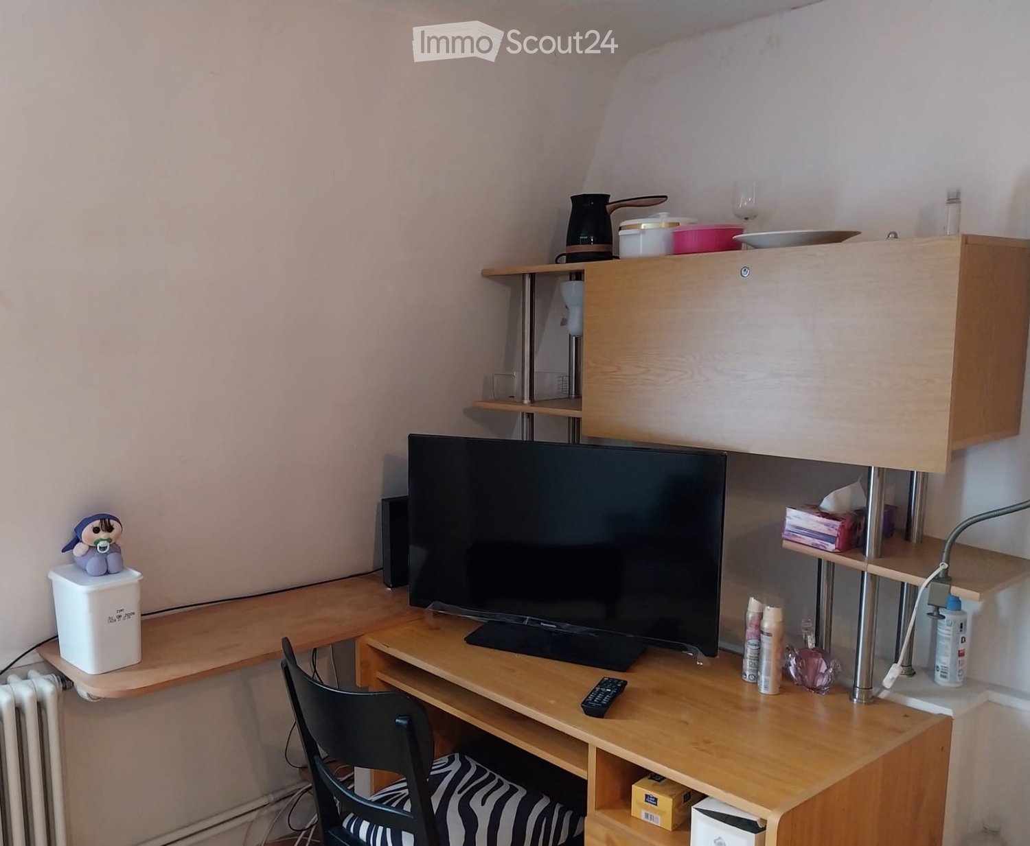 A small, furnished room with a wooden table, a black chair, a large screen, a remote control, a small cabinet, and various items on the shelves and table.