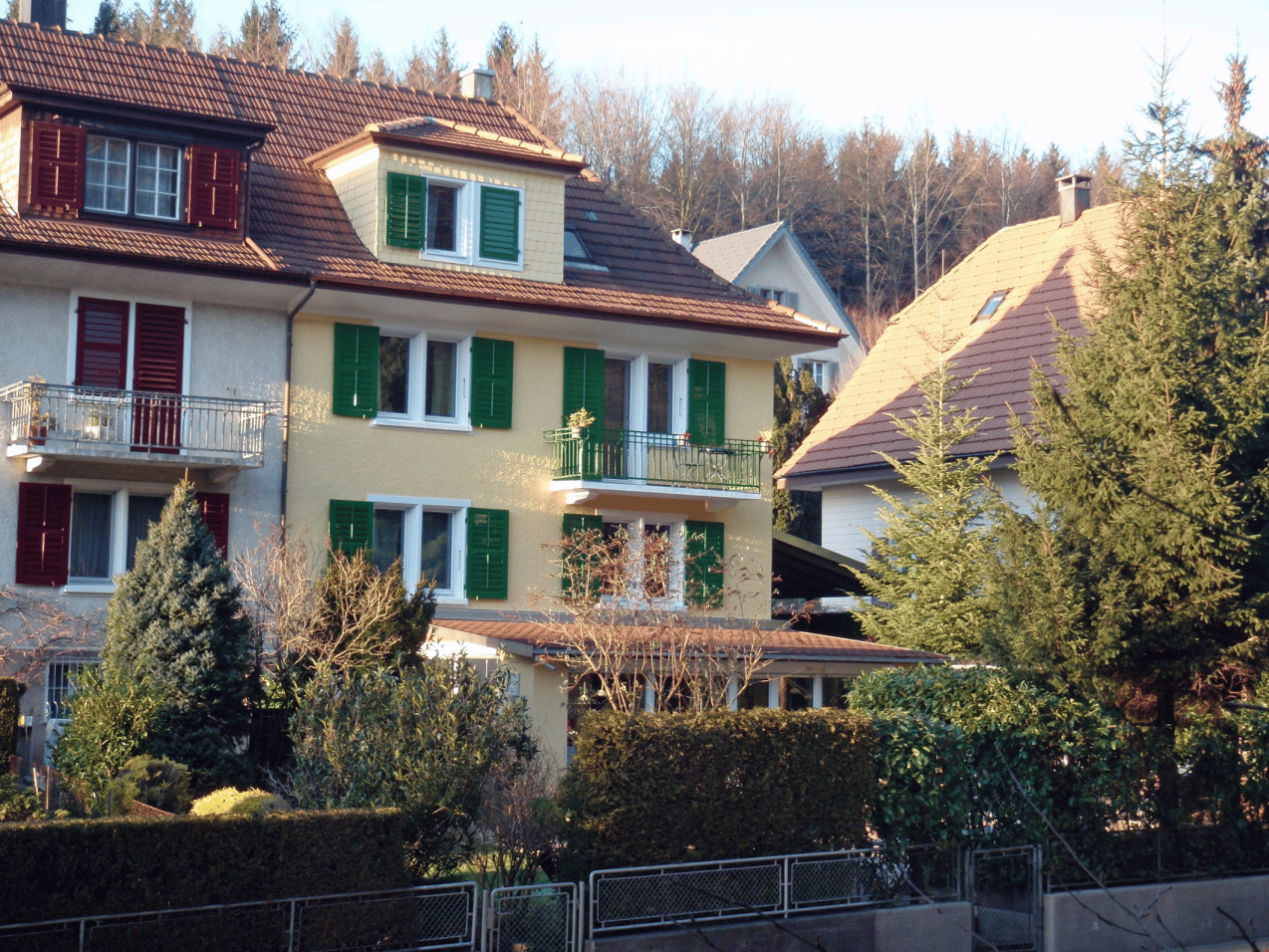 3 story house, yellow exterior, green shutters, brown roof, balcony, garden, trees