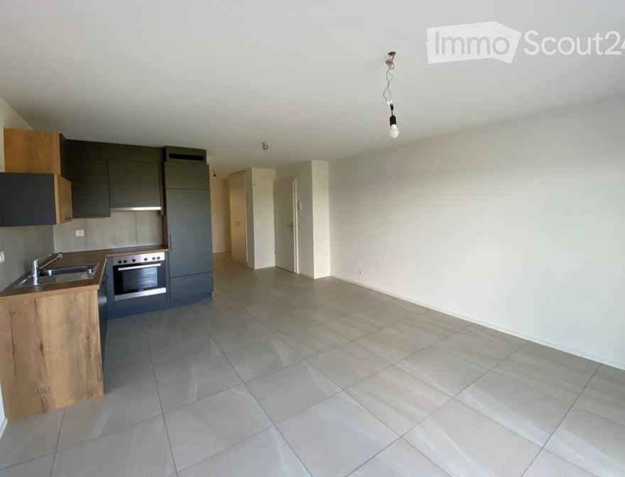 Empty room with tiled floor and white walls, connected to a modern kitchen area with wooden and grey cabinetry, stainless steel sink, and oven.