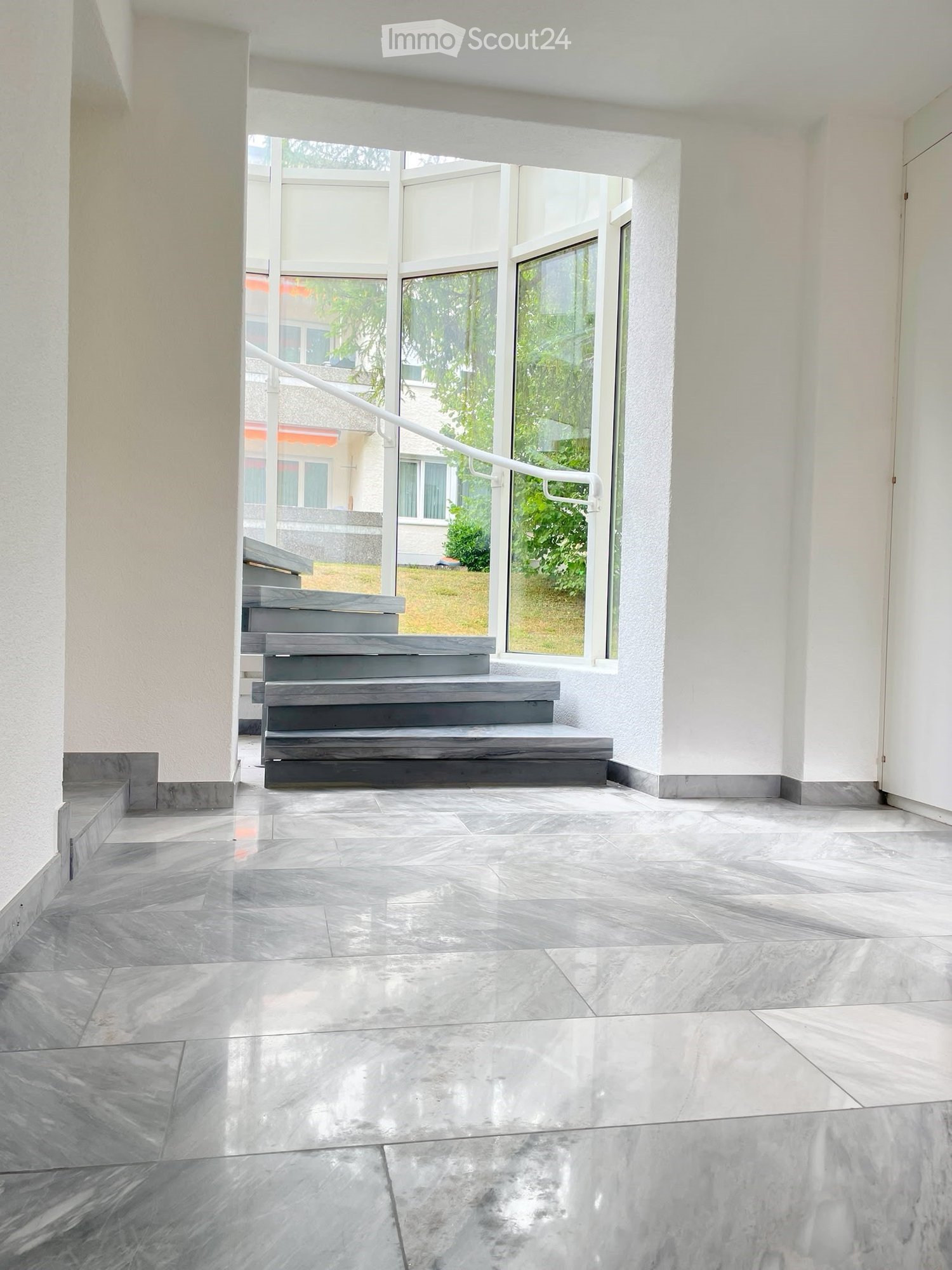 Bright, open hallway with polished marble tiles, white walls, and a staircase leading upwards, featuring large windows that let in natural light.