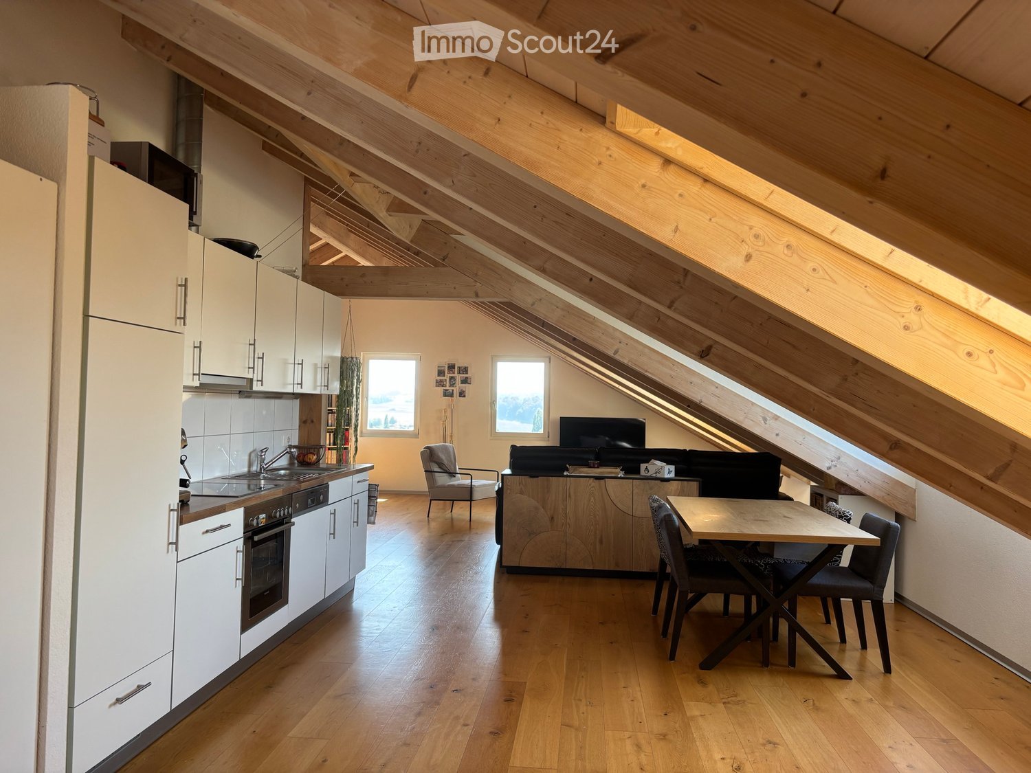 Open-plan living and dining area with exposed wooden beams, modern white kitchen cabinets, and a sleek black sofa.