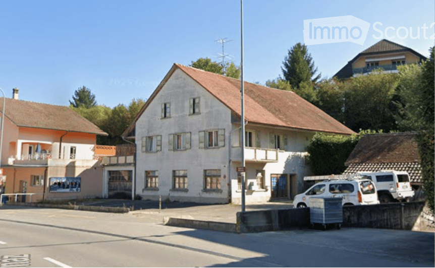 A two-story building with a red-tiled roof and white walls, featuring multiple windows and a small balcony. There are parked vehicles in front, and the building is situated in a residential area with other houses visible in the background.