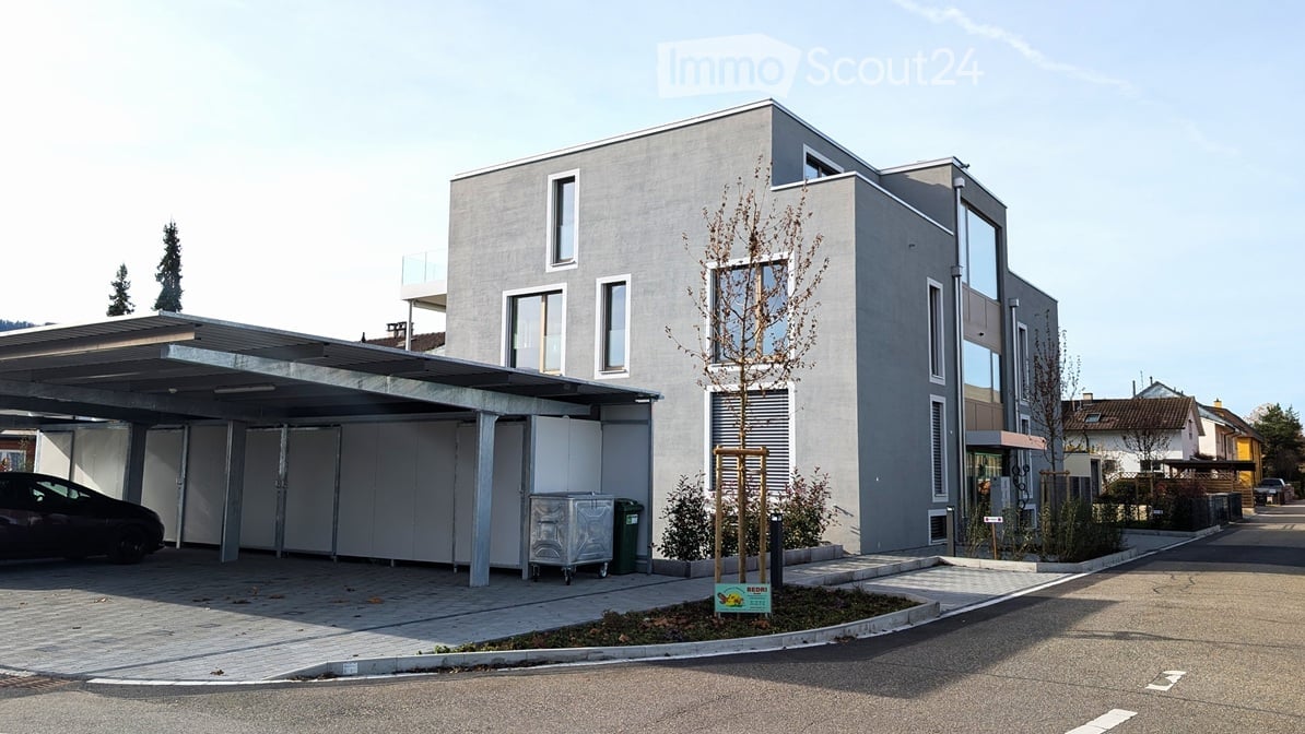 Modern two-story building with a grey facade, large windows, and a covered parking area with multiple car spaces.