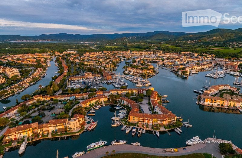 Aerial view of a coastal town with a marina filled with numerous boats and yachts, surrounded by colorful buildings and a scenic backdrop of hills and mountains.