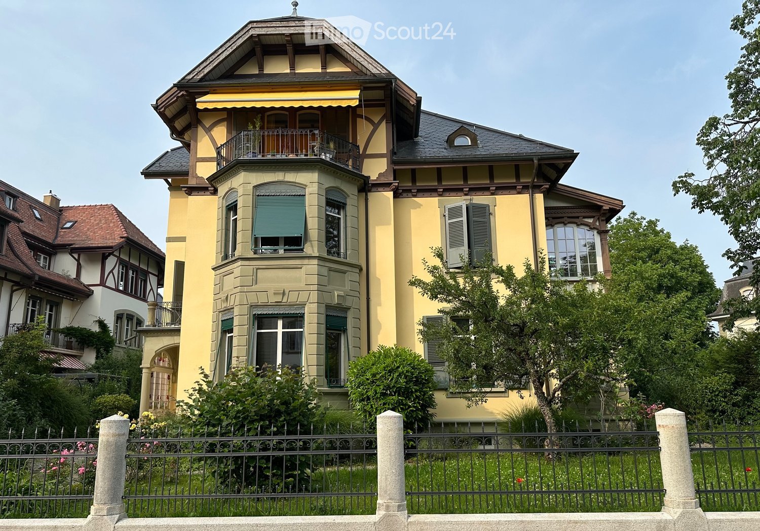 A large, yellow, multi-story house with a prominent balcony, decorative wooden beams, and a well-maintained garden behind a black iron fence.