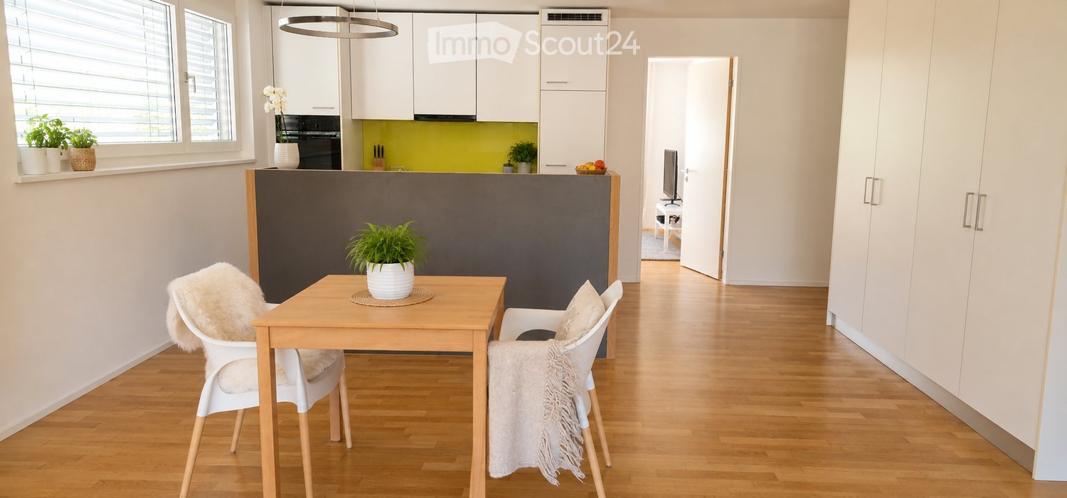 Modern, open-plan living and dining area with a wooden dining table, white chairs, a potted plant, and a kitchen area with white cabinets, an oven, and a refrigerator. The space features light wooden flooring and large windows with blinds.