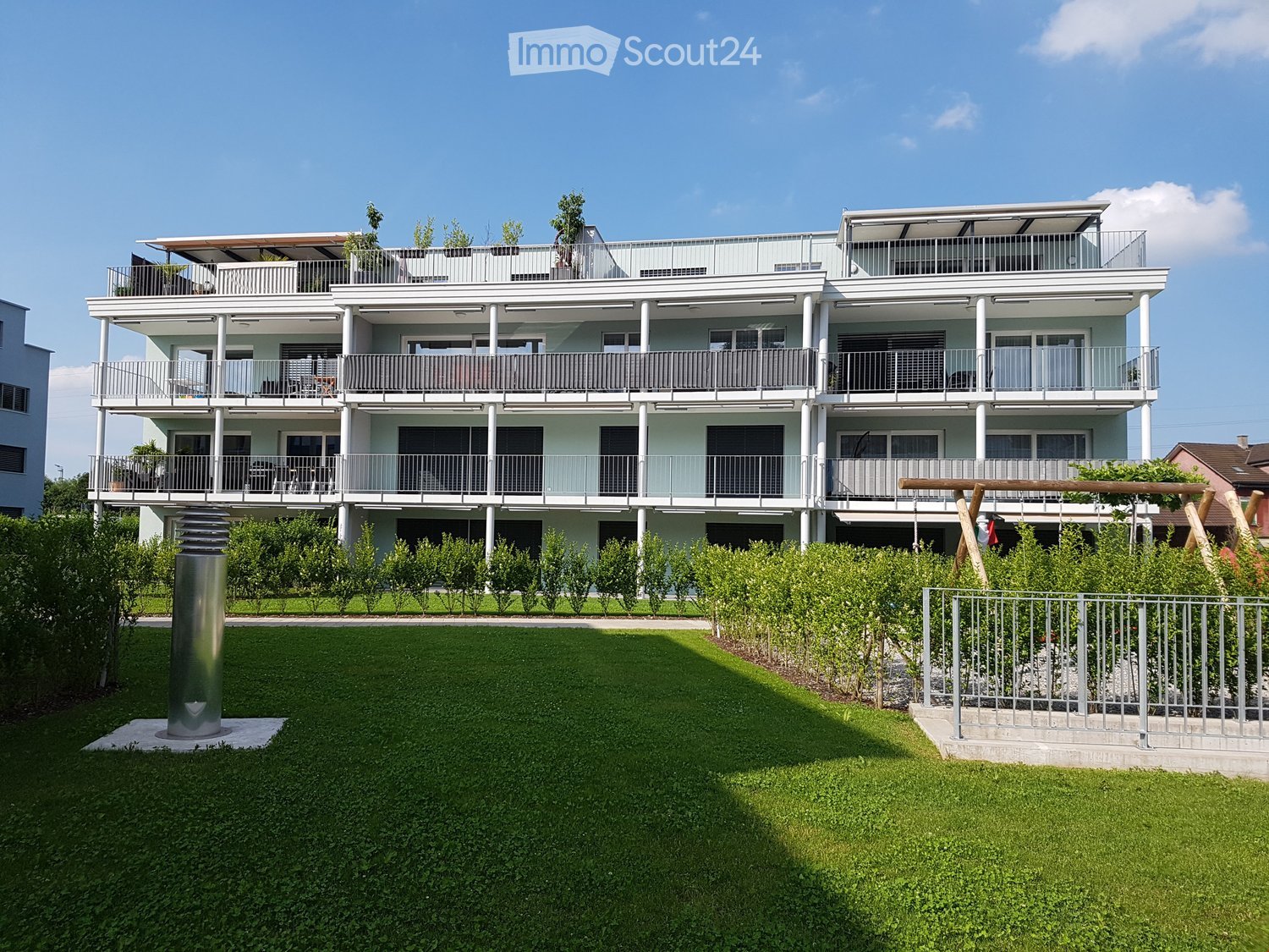 Multi-story apartment building with modern design, large balconies on each floor, green garden area in front, well-maintained surroundings.