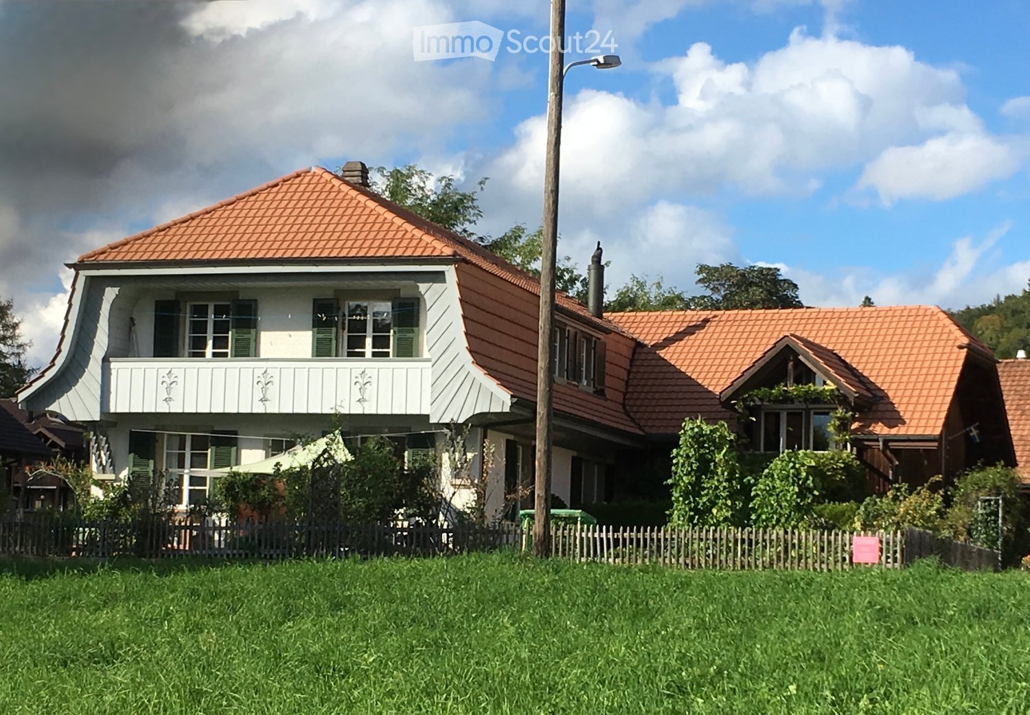 Two-story house with red-tiled roof, white walls, green shutters, and a balcony on the upper floor. The house is surrounded by a wooden fence and greenery.