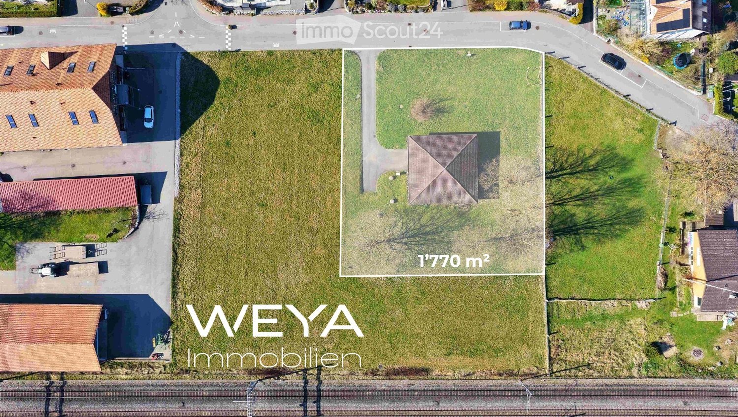 Aerial view of a large, grassy plot of land with a small shed, surrounded by neighboring houses and a road.