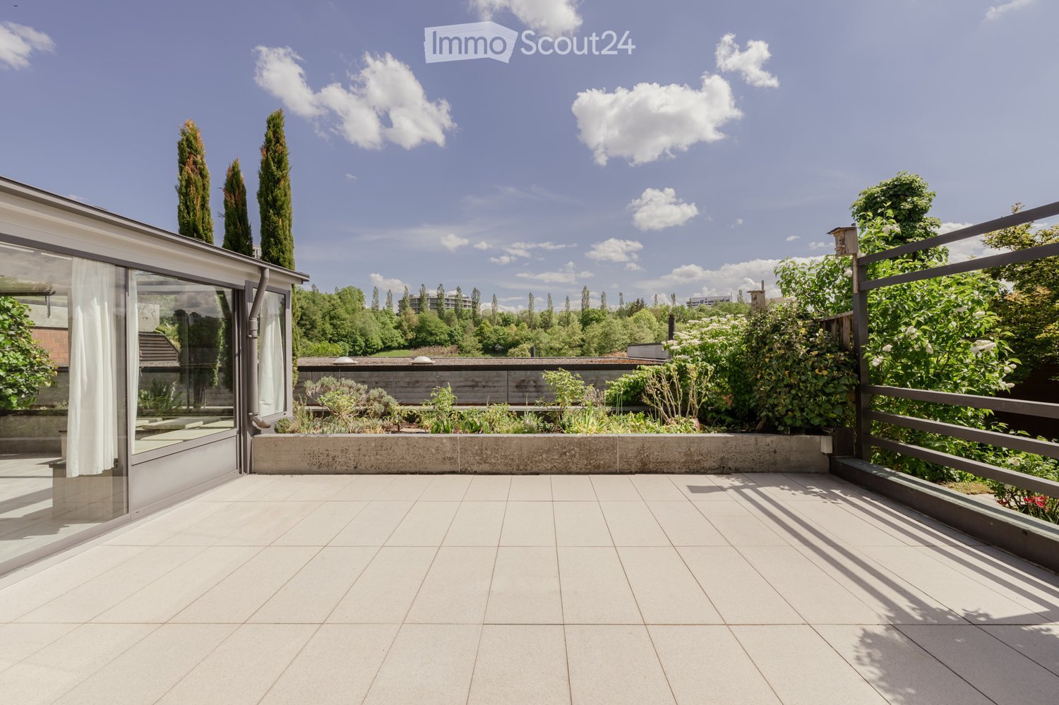 A modern rooftop terrace with white tiled flooring, glass walls, and a metal railing, surrounded by greenery and trees.