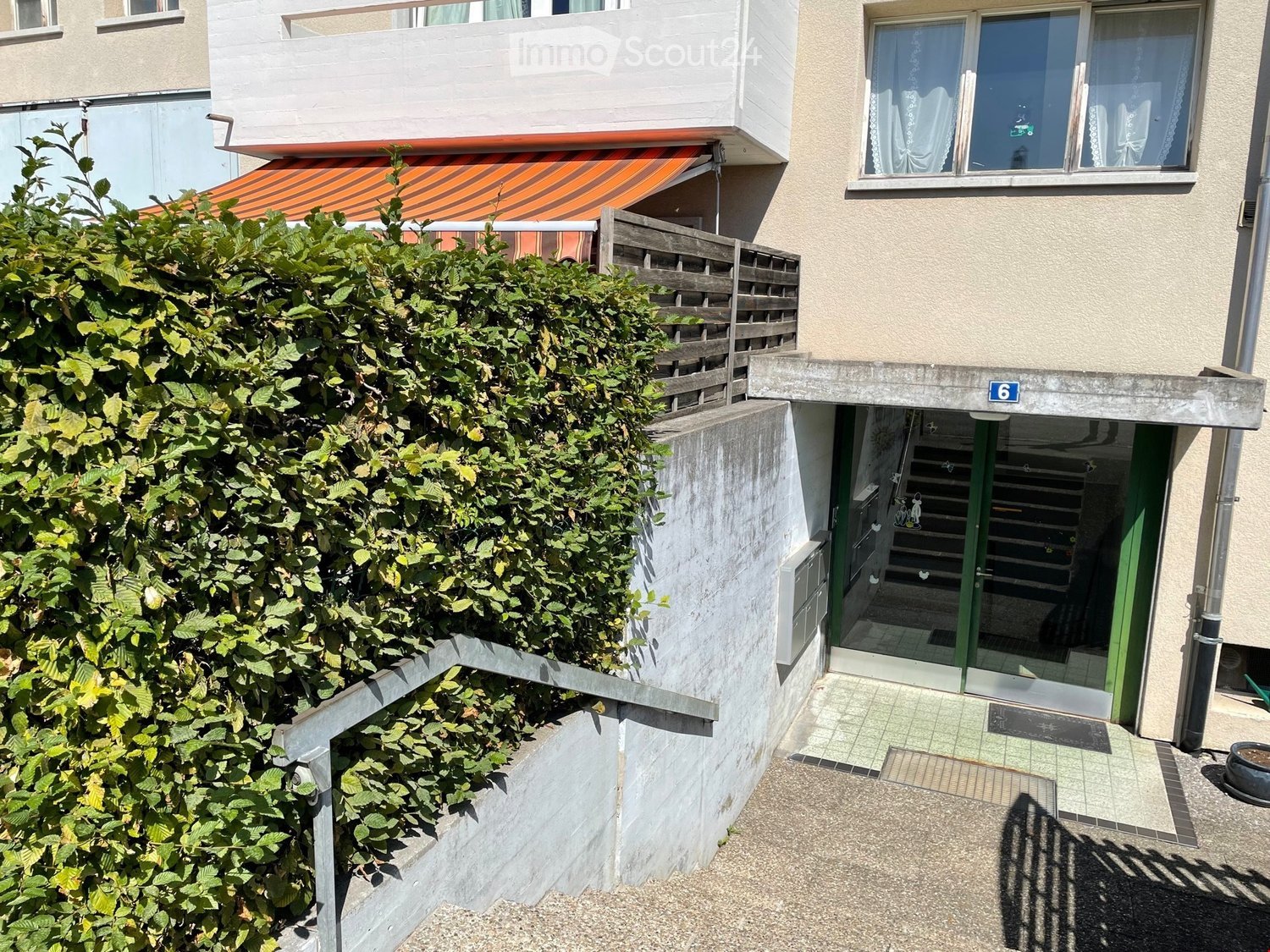 Multi-story residential building with a balcony, covered entrance, and greenery in front.
