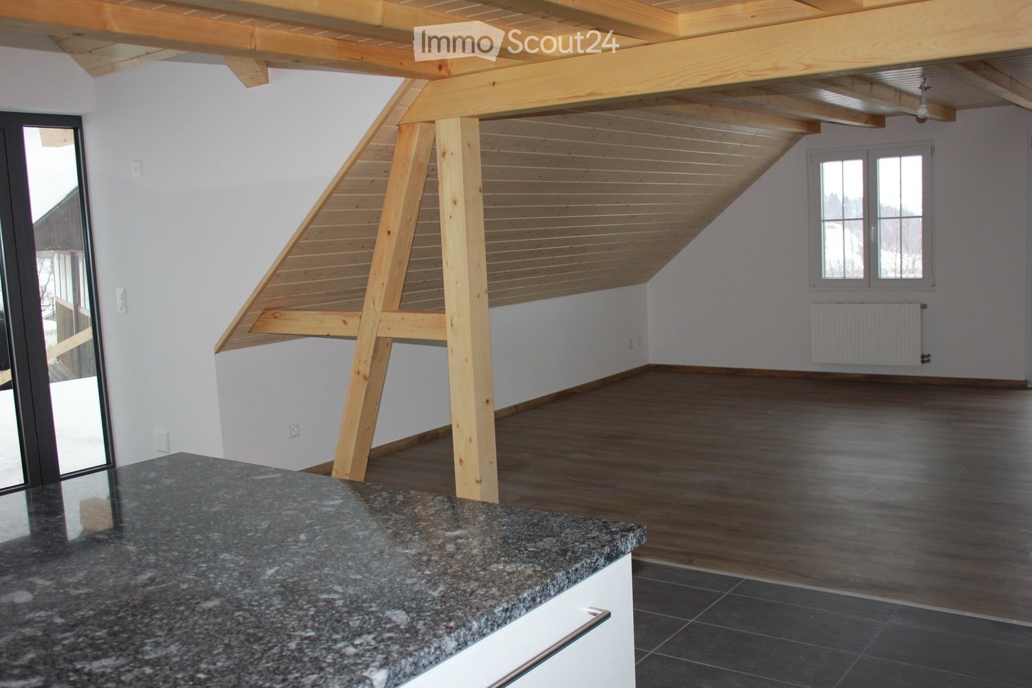 An unfurnished room with wooden beams, a wooden floor, and a granite countertop. The room features large windows and a radiator on the wall.