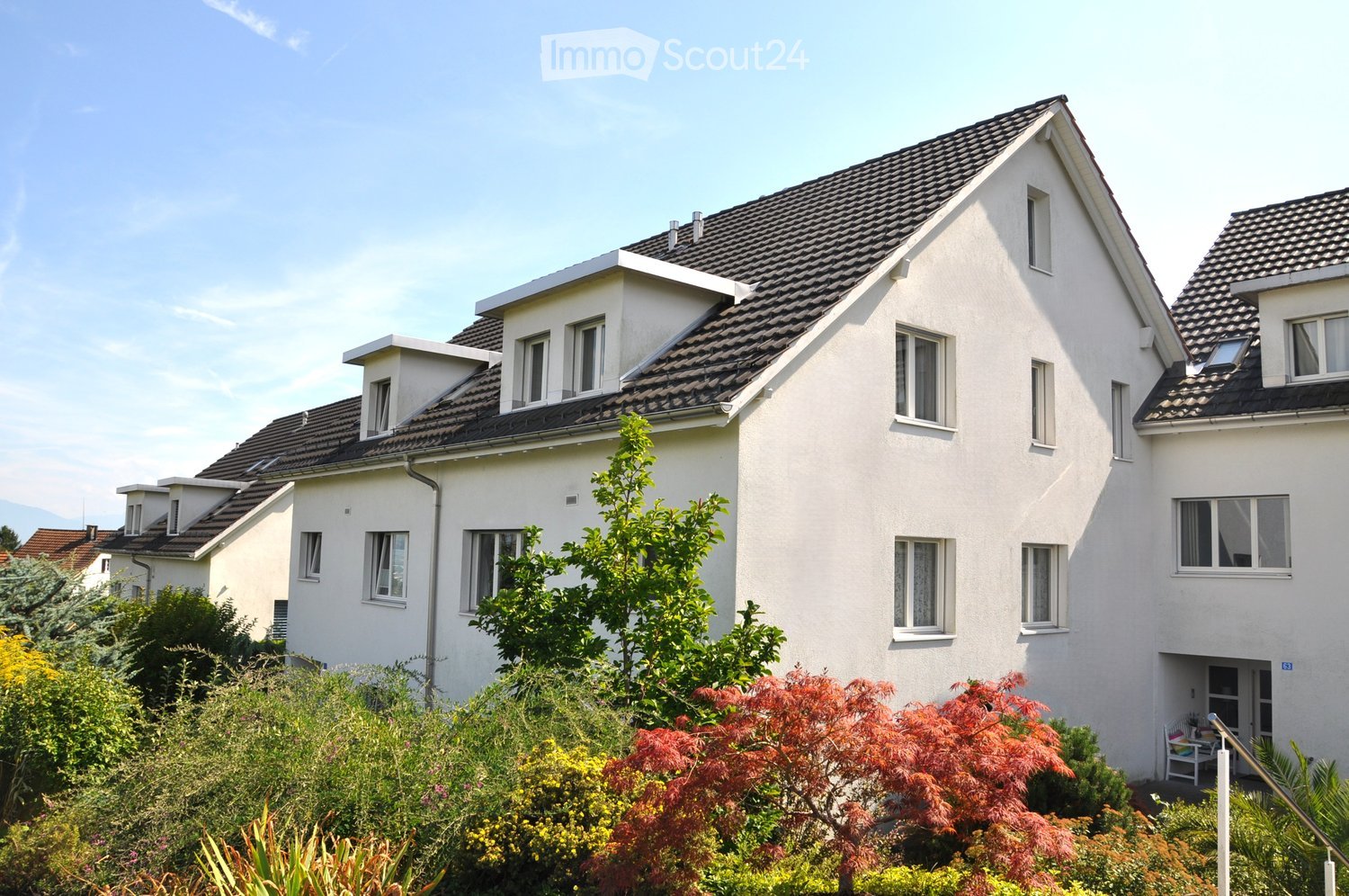 A modern two-story house with white exterior walls, dark grey tiled roof, multiple windows, and a small balcony area visible on the right side.