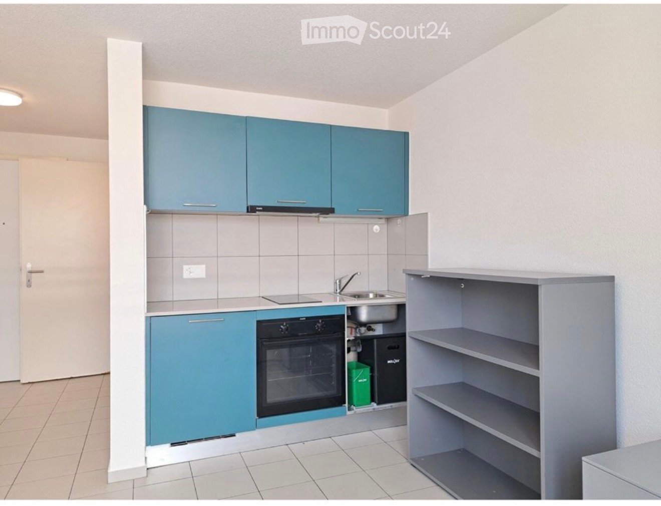 Modern kitchen with blue cabinets, a sink, an oven, and grey shelves. The space is clean and well-organized with tiled walls and a tiled floor.