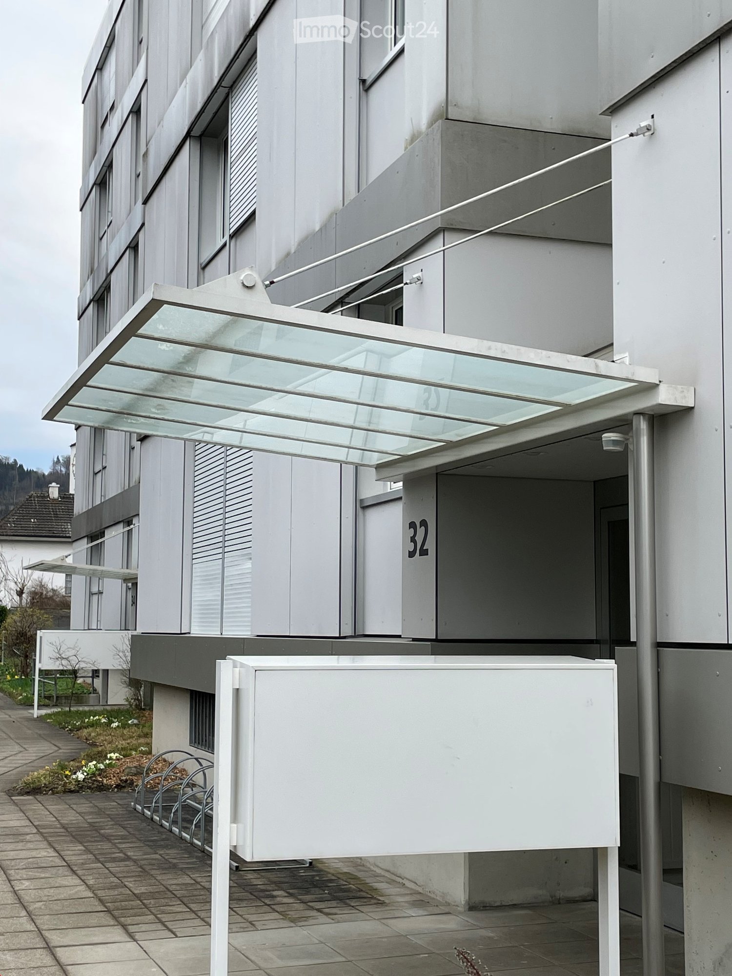 Modern multi-story building with a glass-covered entrance canopy and a white signpost displaying the number 32.