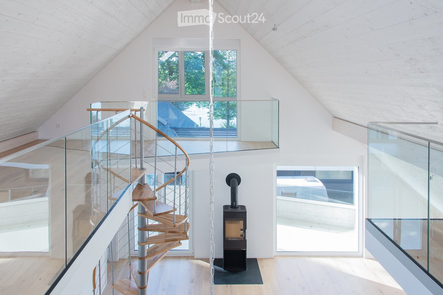 Modern, open-concept hallway with a wooden spiral staircase, glass railings, and a wood-burning stove. Large windows provide natural light and views of the outdoors.