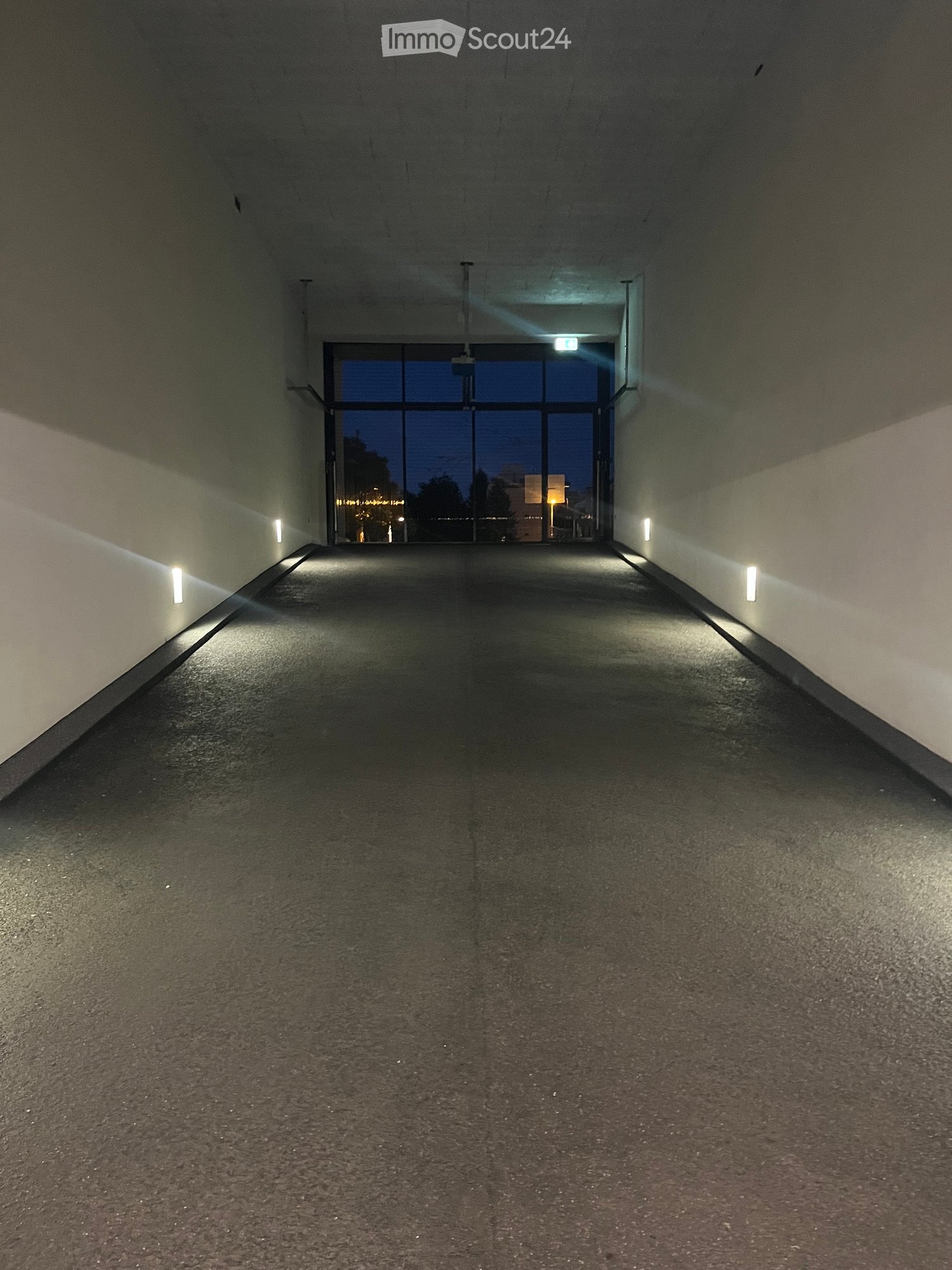 Empty hallway with concrete walls, carpeted floor, and recessed lighting, leading to a glass door opening to the outside.