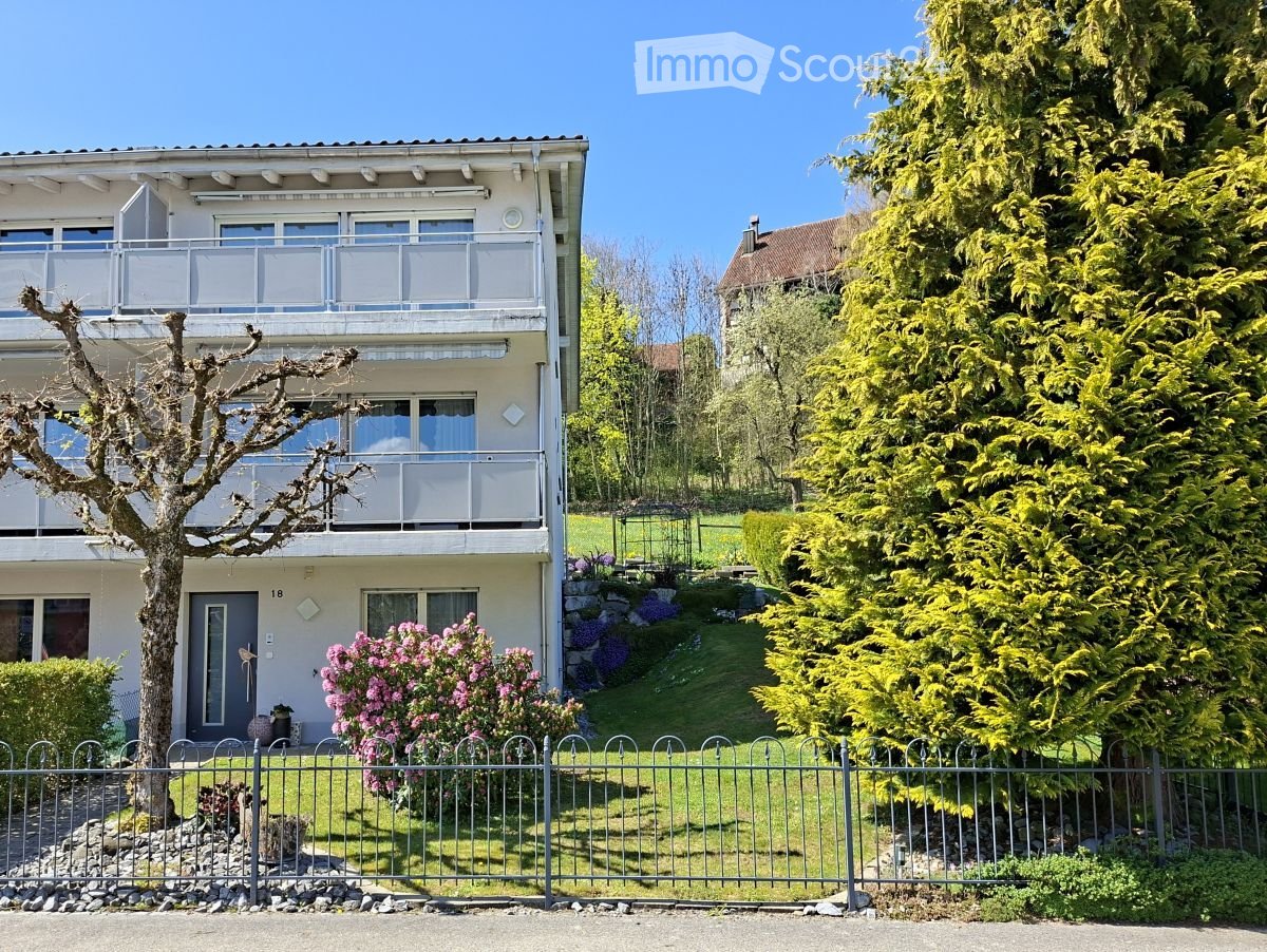 Two-story house with balconies, surrounded by a well-maintained garden with trees and flowering shrubs.