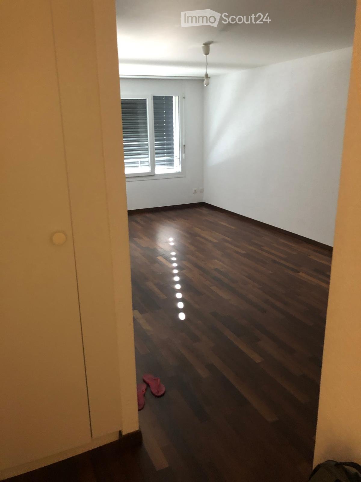 Empty room with wooden flooring, white walls, and a window with blinds. A pair of pink slippers is placed near the window.