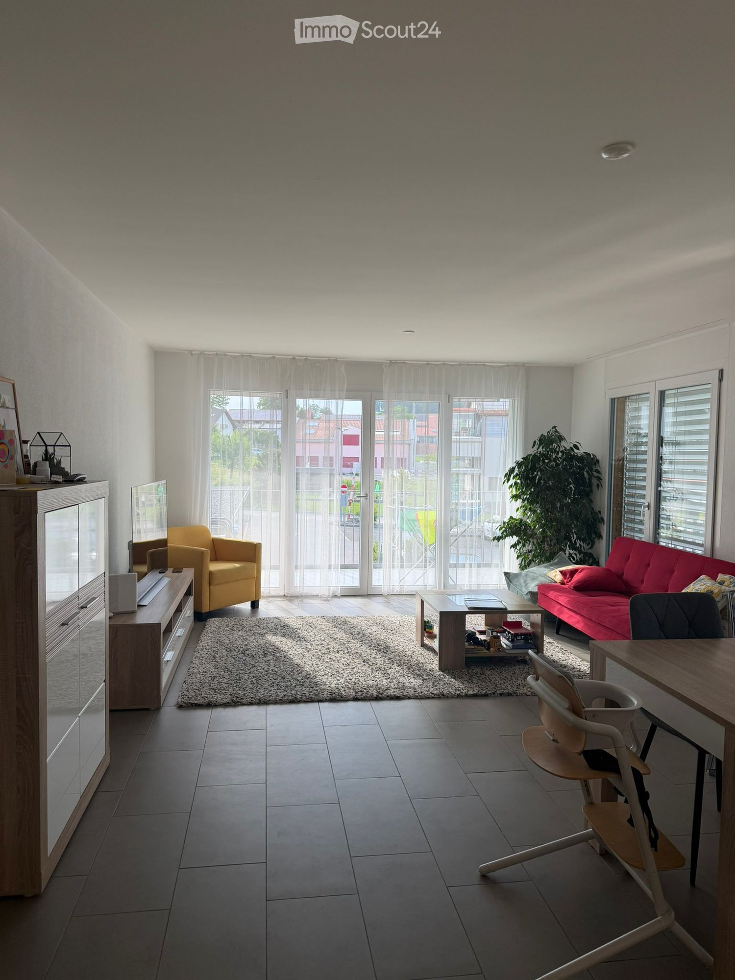Modern living room with large windows and sliding doors leading to a balcony. Features a red sofa, yellow armchair, coffee table, dining table with chairs, and a wooden cabinet with a TV.