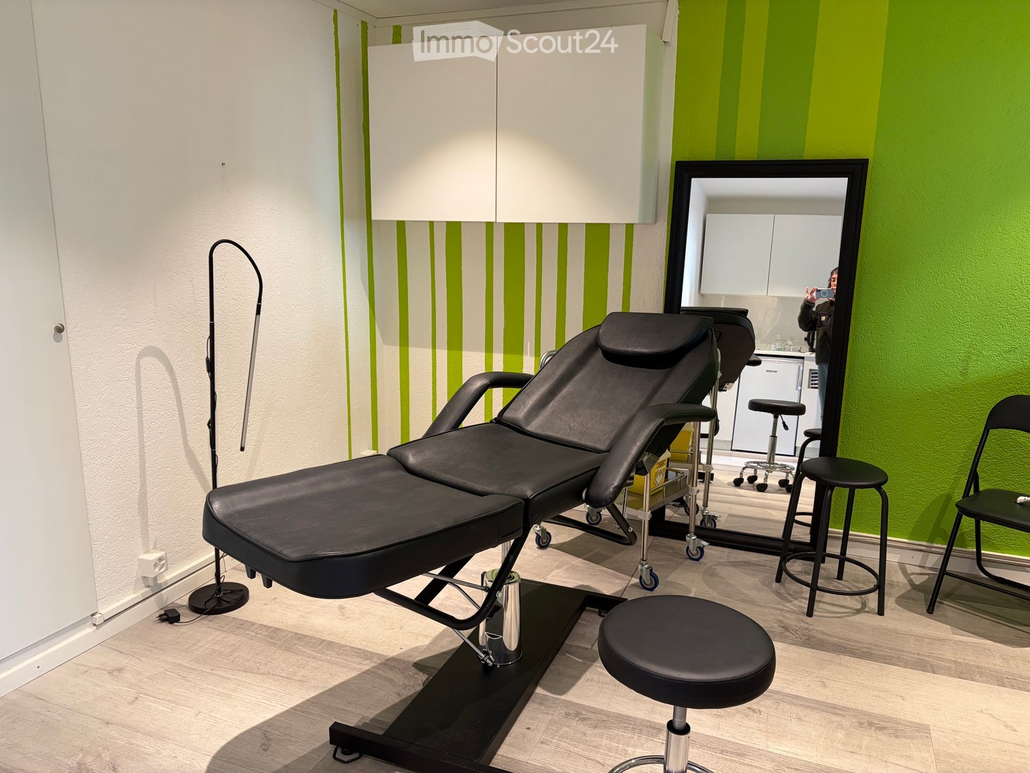 Modern massage room with black leather massage chair, adjustable stools, large mirror, white cabinet, and sleek flooring.