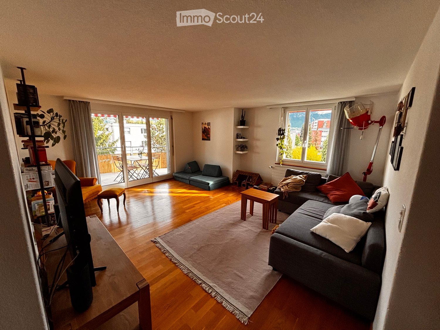 Bright living room with wooden flooring, large windows, and a view. Features a grey sectional sofa, coffee table, small table, and a TV on a stand.