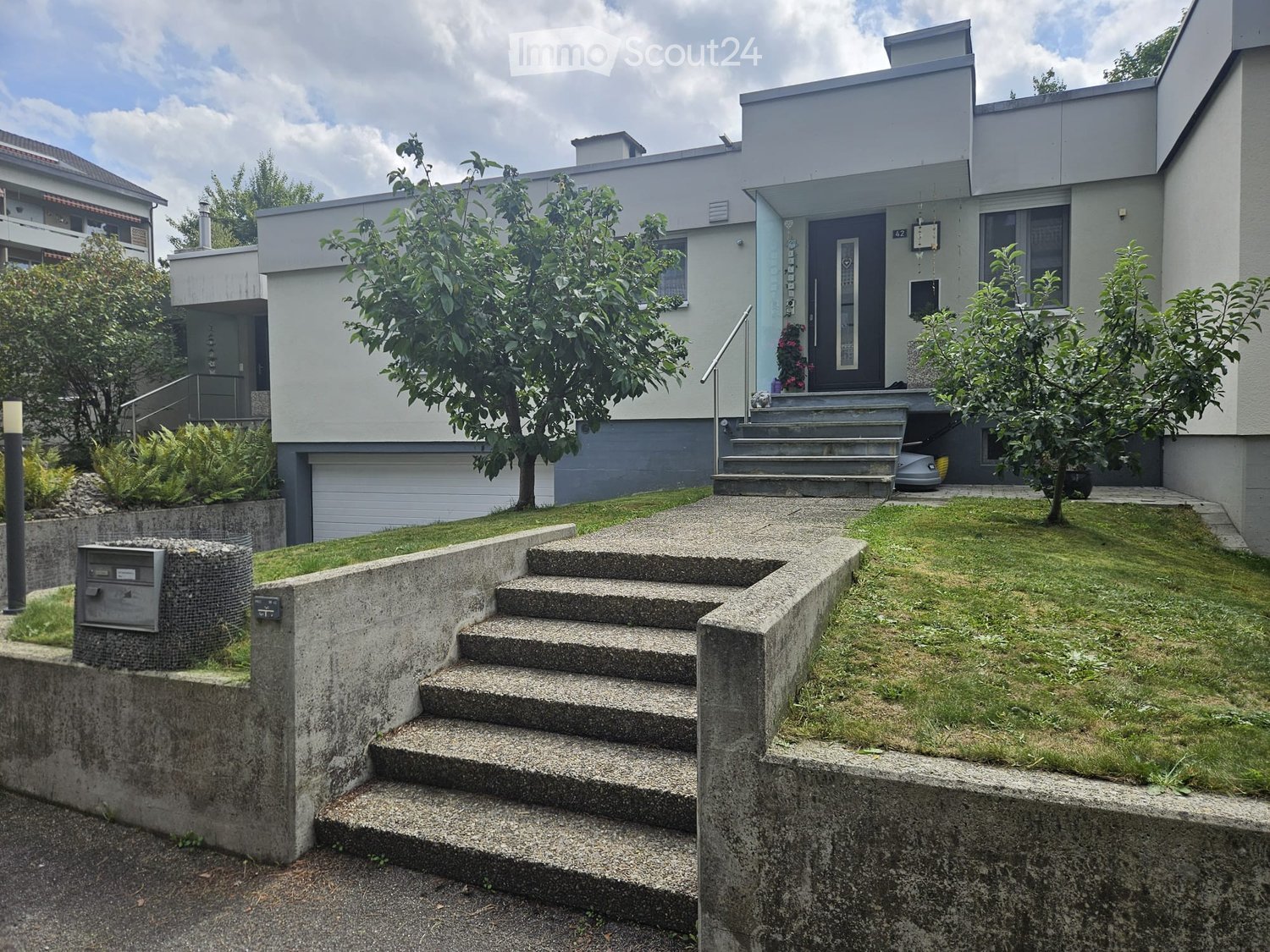 Modern two-story house with a raised ground floor, concrete steps leading to the entrance, and a small garden area with trees and shrubs.