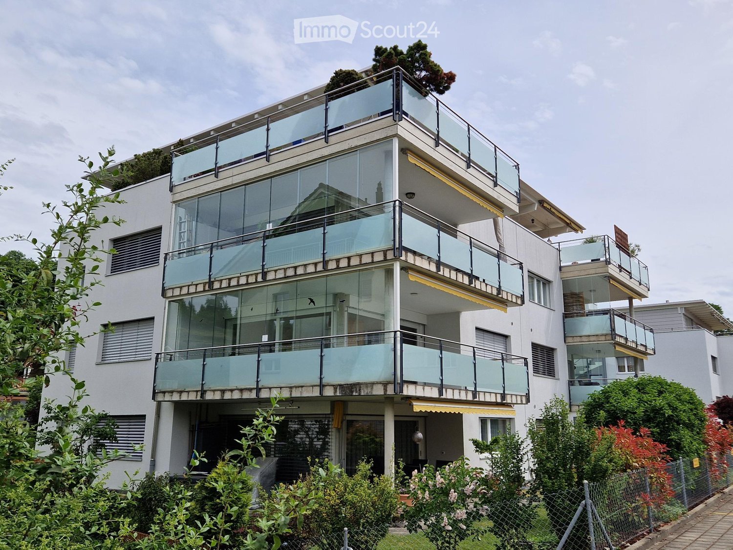 Modern multi-story building with large glass balconies and contemporary design, surrounded by greenery.