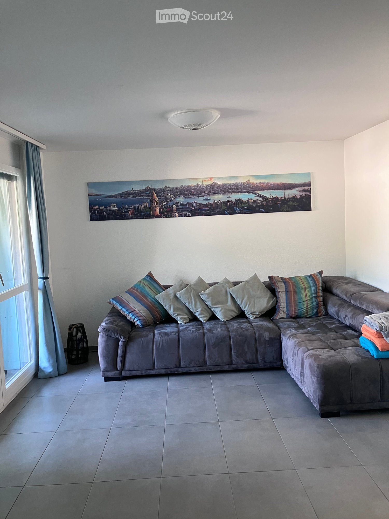 Modern living room with a large sectional sofa, colorful pillows, a large cityscape picture on the wall, and a window with curtains.