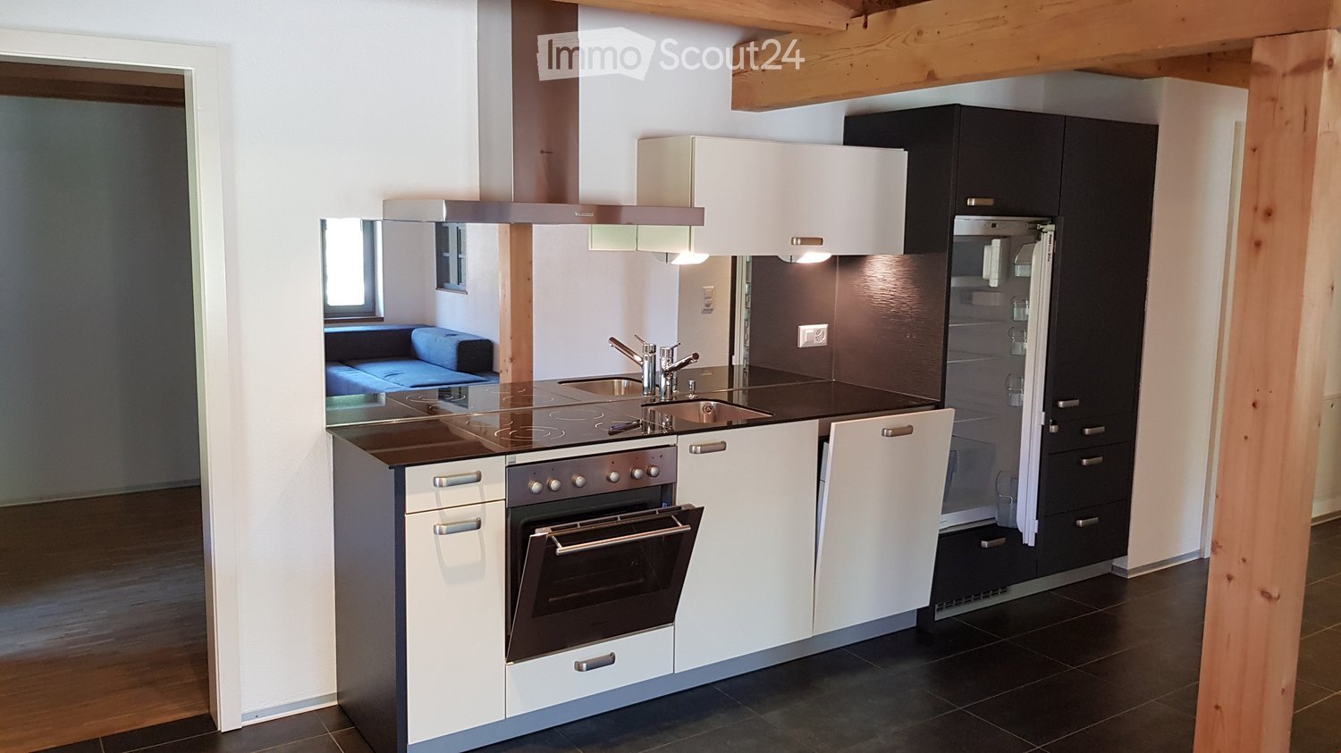 Modern kitchen with white cabinets, black appliances including oven and dishwasher, wooden beams on the ceiling, and a blue sofa visible in the adjacent room.