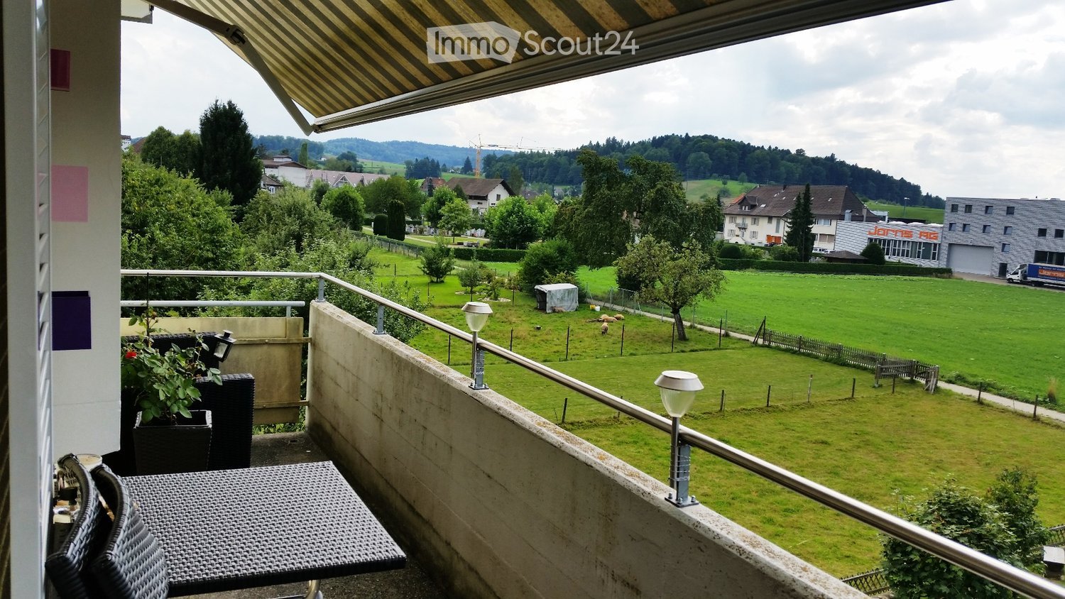 Balcony with table and chairs, metal railing, retractable awning, view of a grassy field with trees and houses