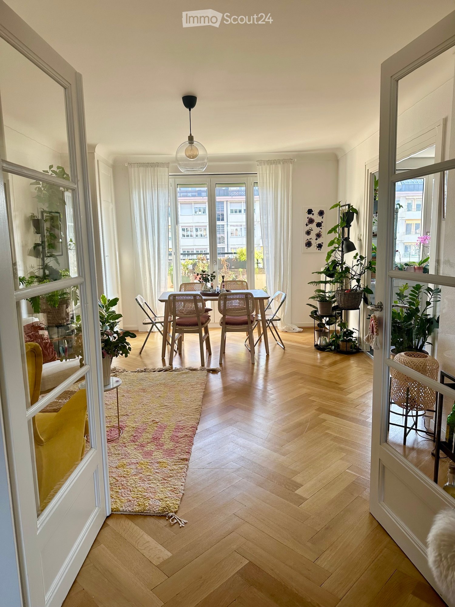 Bright living room with wooden flooring, a yellow sofa, a dining table with chairs, a colorful rug, and several potted plants near the windows and balcony.