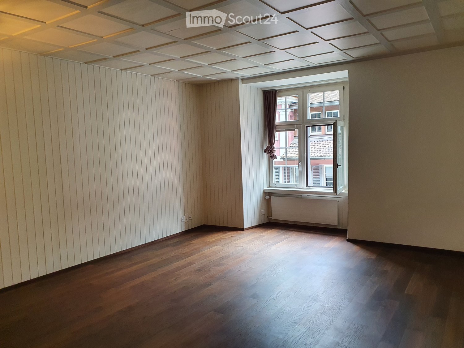 Empty room with wooden flooring, white paneled walls, and a large window with a curtain. The ceiling features a decorative coffered design.